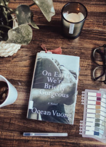 A flatlay photo of the book On Earth We're Briefly Gorgeous by Ocean Vuong on a dark surface. Surrounding the book on the surface are some reading tabs, a cup of tea, a pen, glasses, a candle, and a housepland.