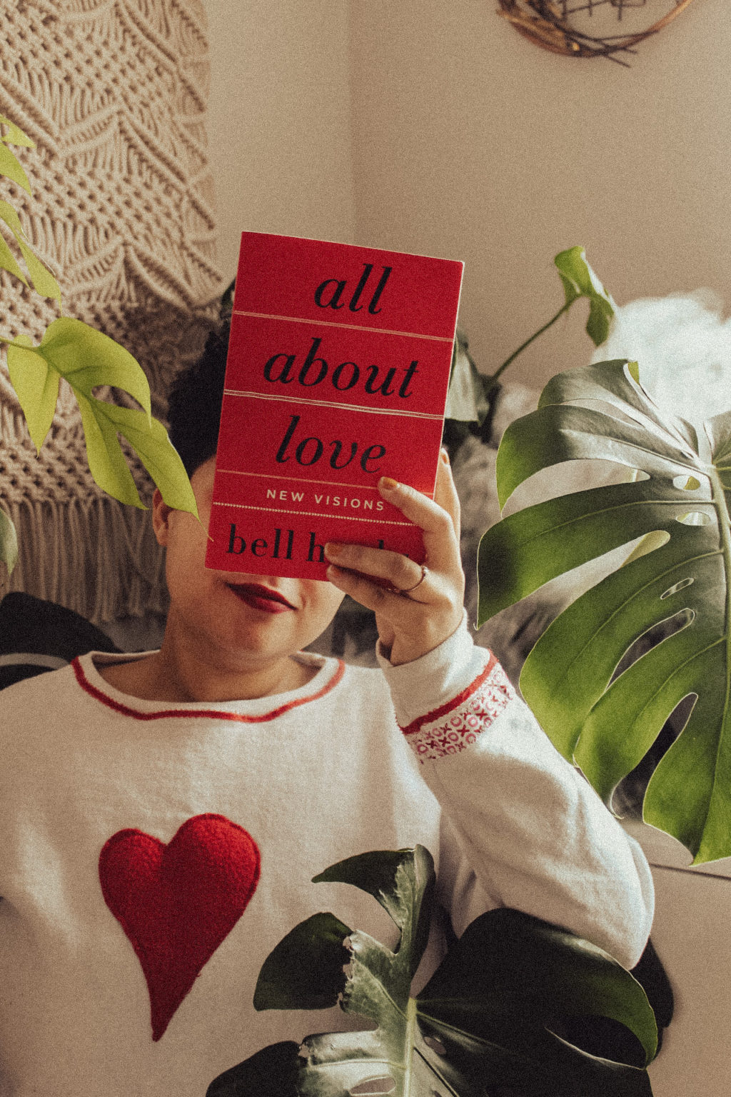 Emily (the author) holding up a copy of All About Love by Bell Hooks in front of her face, wearing a white sweater with a red heart on it. She is surrounded by some large leaves from her monstera deliciosa plants.