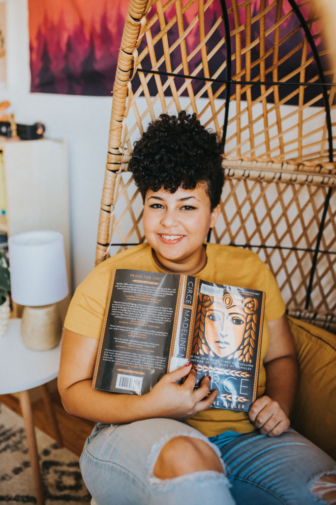 Emily, the owner of the bog, sitting in an egg chair with the book Circe by Madeline Miller open in front of her. She is wearing a yellow shirt and ripped jeans.