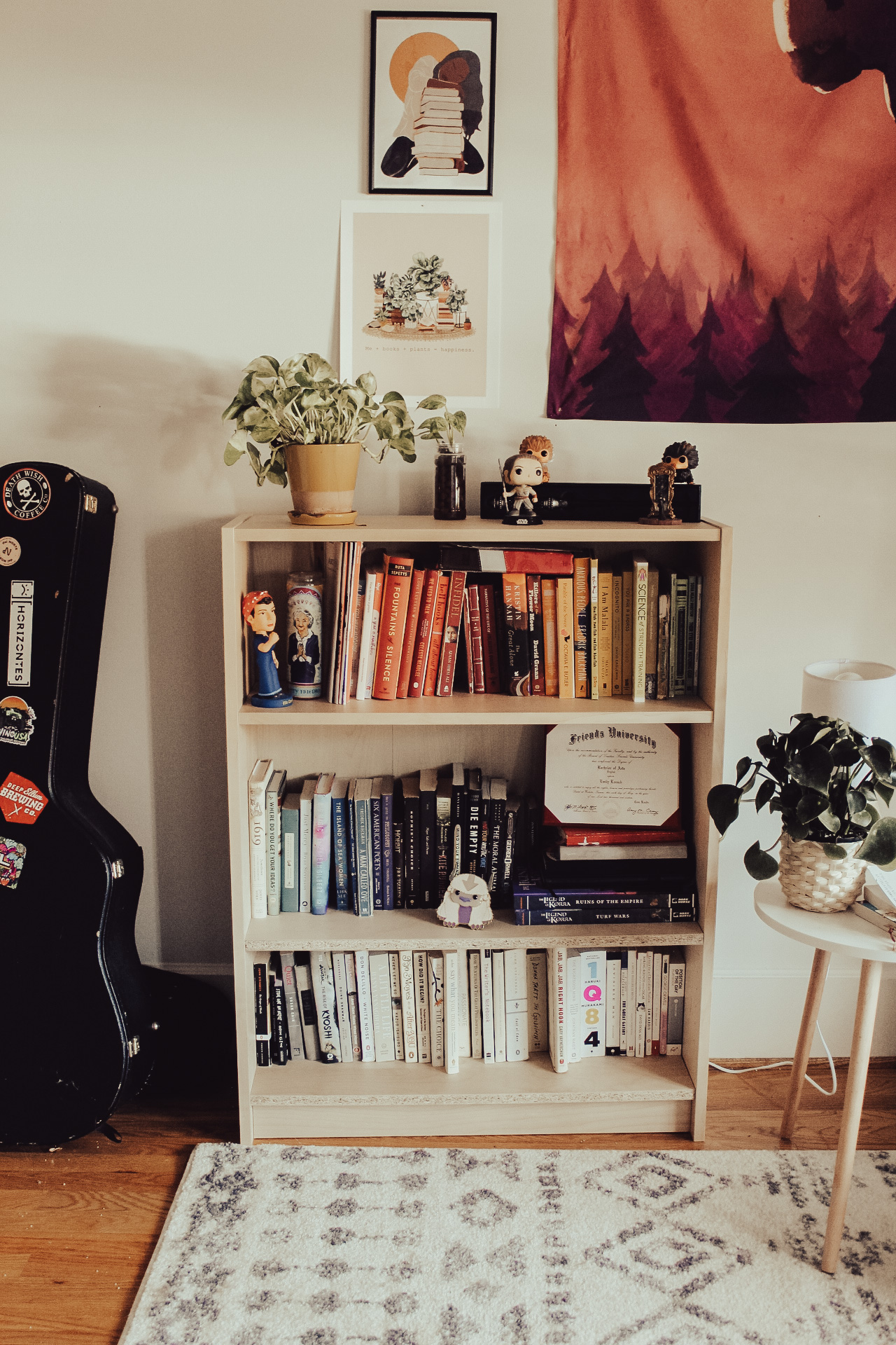 An IKEA bookshelf with book organized by color on it. Two plants are sitting on top of the shelf as well as some Harry Potter vinyl figures. A guitar case rests next to the bookshelf, and some artwork including an Avatar tapestry are hung on the wall above the shelf.