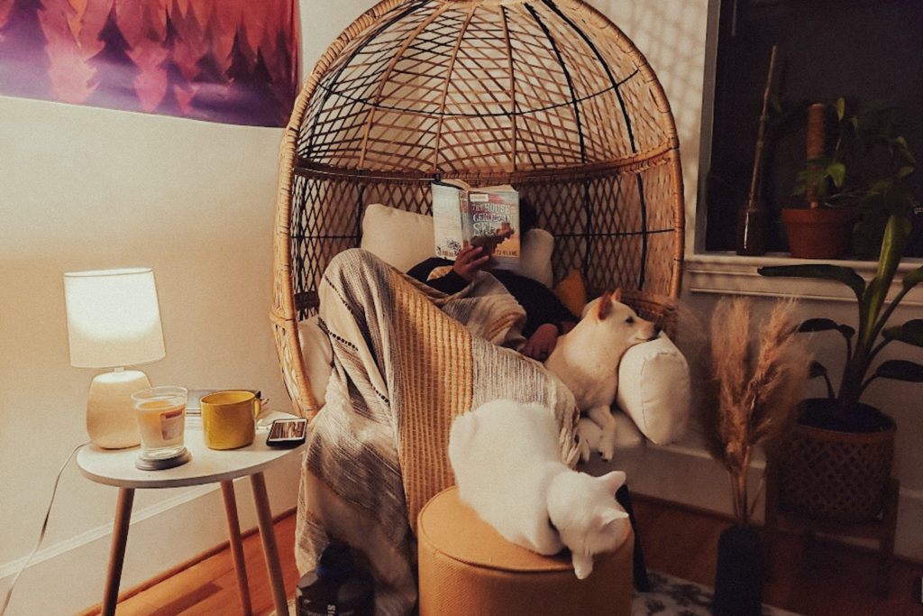 Emily is sitting inside the egg chair in her reading nook reading the book The House in the Cerulean Sea by T.J. Klune. Her shiba inu is snuggled on the chair with her while her cat Olaf rest on the footrest.