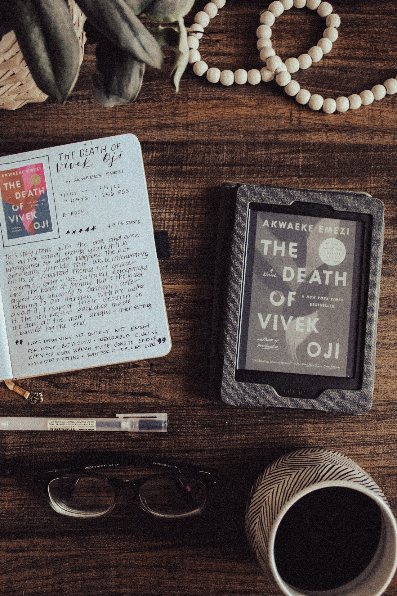 A book journal set up with a book review of The Death of Vivek Oji by Akwaeke Emezi. Journal page is next to a Kindle Paperwhite with the book displayed on the screen. Glasses, pen, mug, and plant scattered around photo.