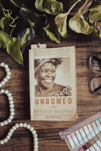 A flatlay photo of the book Unbowed by Wangari Maathai displayed on a dark table. Wood beads, reading tabs, glasses, and plants surround the surface around the book.