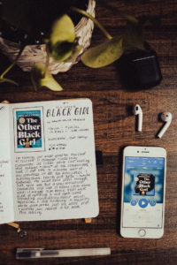A book journal set up with a book review of The Other Black Girl by Zakiya Delilah Harris. Journal page is next to an iPhone with the audiobook version of the book displayed, a plant, and a set of Apple Airpods.