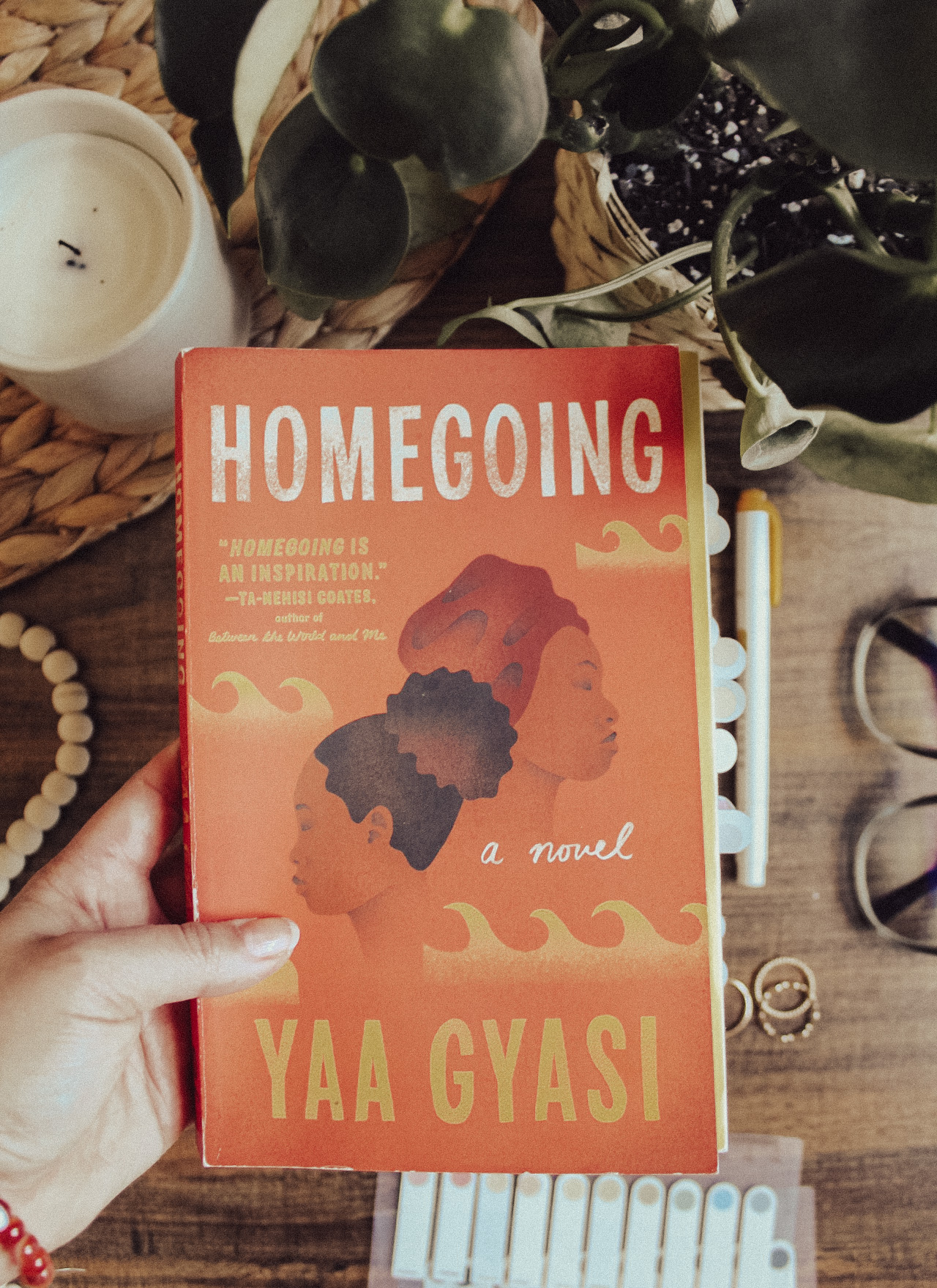 Flatlay photo of the book Homeoging by Yaa Gyasi with a dark background. Surface is surrounded with a candle, glasses, reading tabs, and a houseplant.