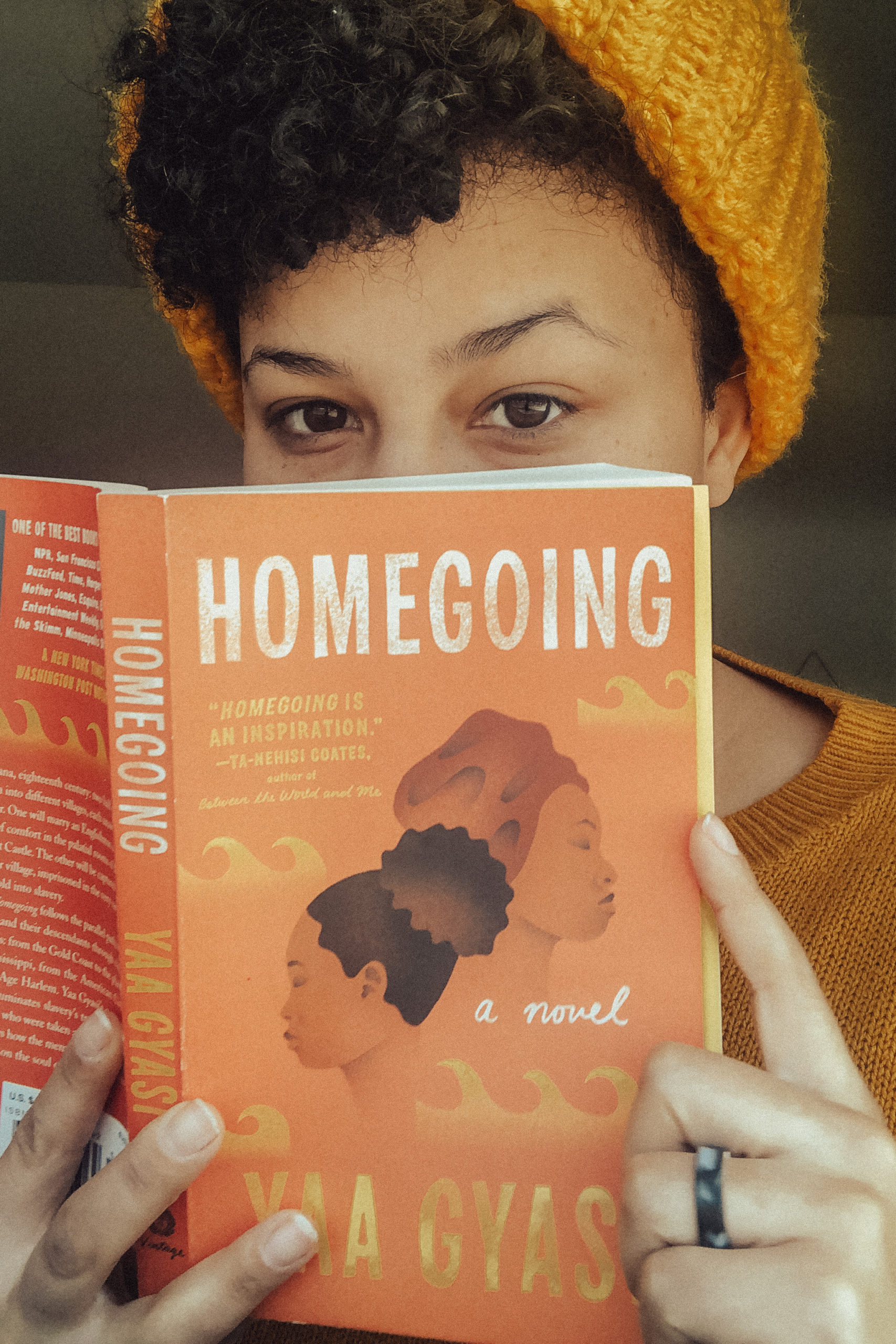 Photo of me, Emily, wearing a yellow beanie and holding the book Homegoing by Yaa Gyasi in front of my face. The book cover is red, with two Black women displayed on the front.