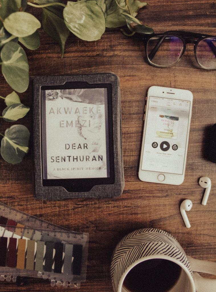 A flatlay photo of a Paperwhite Kindle with Dear Senthuran by Akwaeke Emezi on a tabletop background. Next to the e-book is an audiobook version of the memoir, surrounded by houseplants. airpods, glasses, a cup of coffee, and some reading tabs.