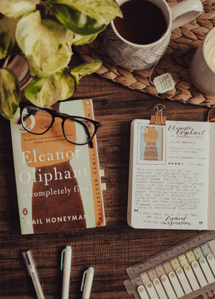 Flatlay photo of a paperback copy of the book Eleanor Oliphant is Completely Fine by Gail Honeyman. Next to the book is a reading journal entry and book review for the story. Decorative elements such as reading tabs, plants, glasses, and a cup of coffee are around the book and book journal.