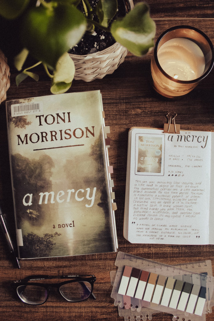 A flatlay photo of the hardback version of A Mercy by Toni Morrison. Next to the book is my book bullet journal with my book review entry for A Mercy in it. Decorative elements surround the book and journal, including a houseplant, reading tabs, glasses, and a candle.