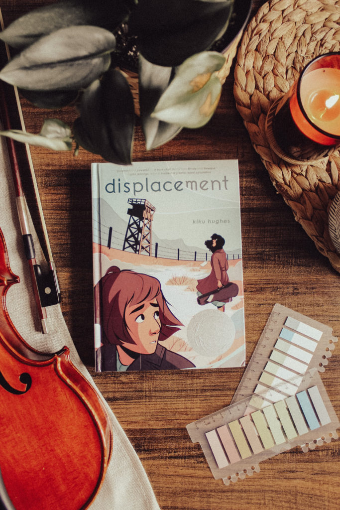 The young adult graphic novel Displacement by Kiku Hughes is laying on a dark tabletop background, surrounded by decorative elements such as reading tabs, a viola and bow, a houseplant, and a candle.