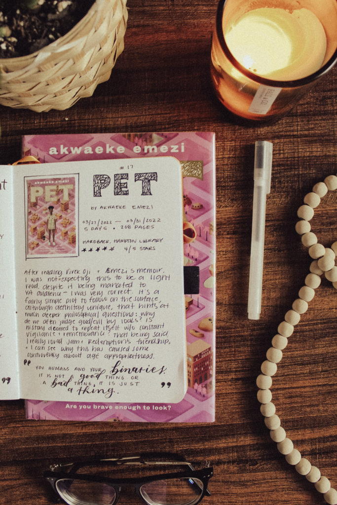 A book journal entry for the young adult novel Pet by Akwaeke Emezi is lying open on a hardback copy of the book. Decorative elements surround it, such as a pen, wood beads, a candle, and a houseplant, all on a dark tabletop background.