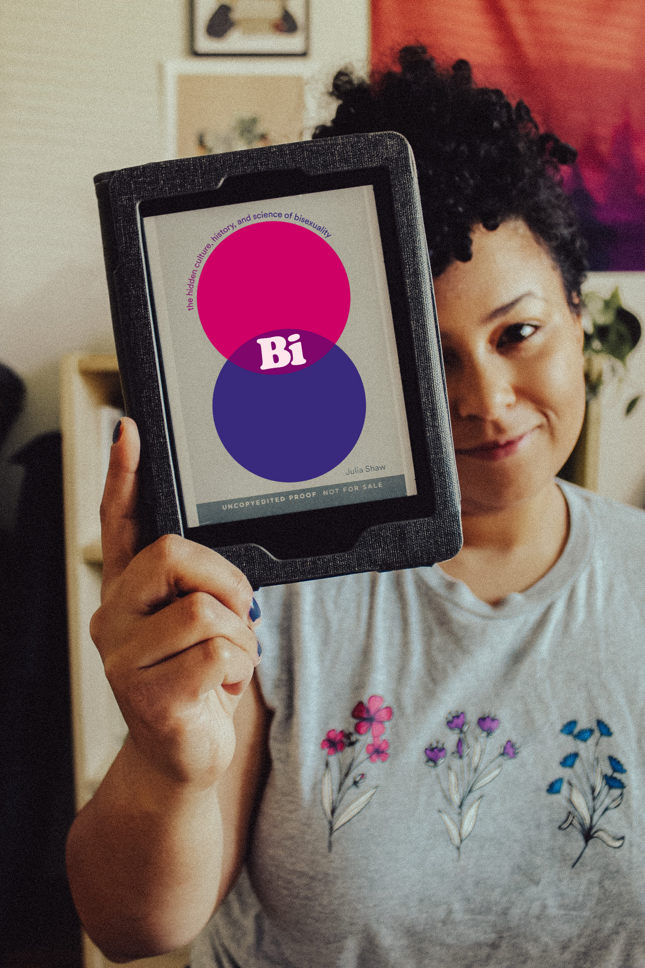 Emily holding up an e-book copy of Bi: The Hidden Culture, History, and Science of Bisexuality by Julia Shaw. Wearing a shirt with the bisexual pride colors for pride month.