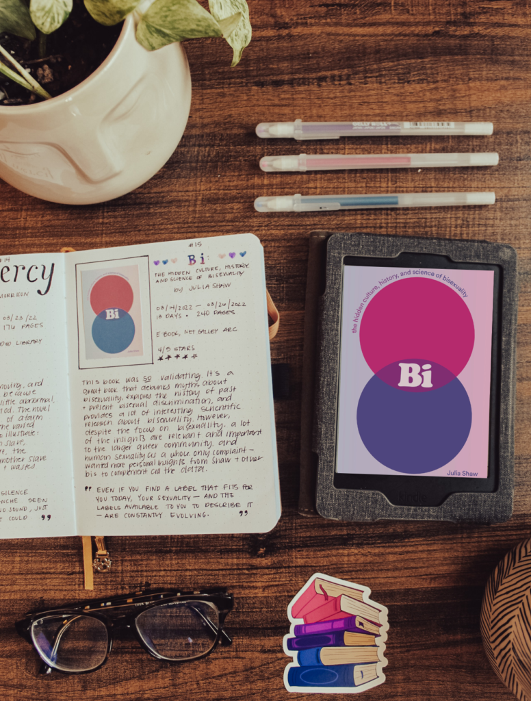 Flatlay photo of the e-book version of Bi: The Hidden Culture, History, and Science of Bisexuality by Julia Shaw. Next to the book is a book journal entry for the book. Surrounded by bi pride colors, glasses, a candle, and a houseplant.