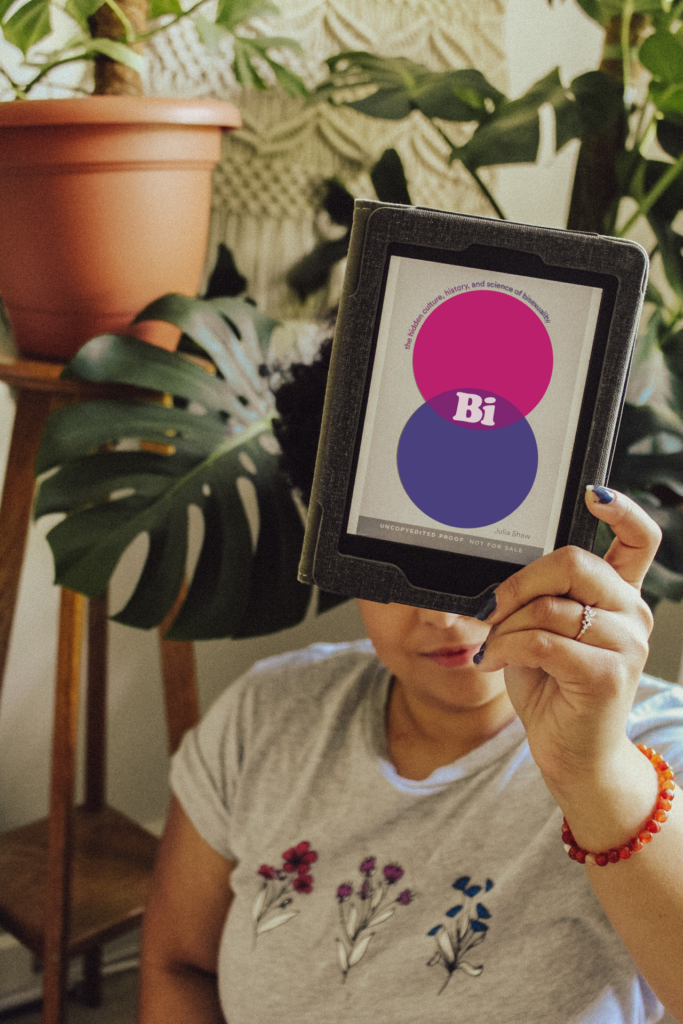 Emily holding up an e-book copy of Bi: The Hidden Culture, History, and Science of Bisexuality by Julia Shaw. Wearing a shirt with the bisexual pride colors for pride month. Green florals in the background.