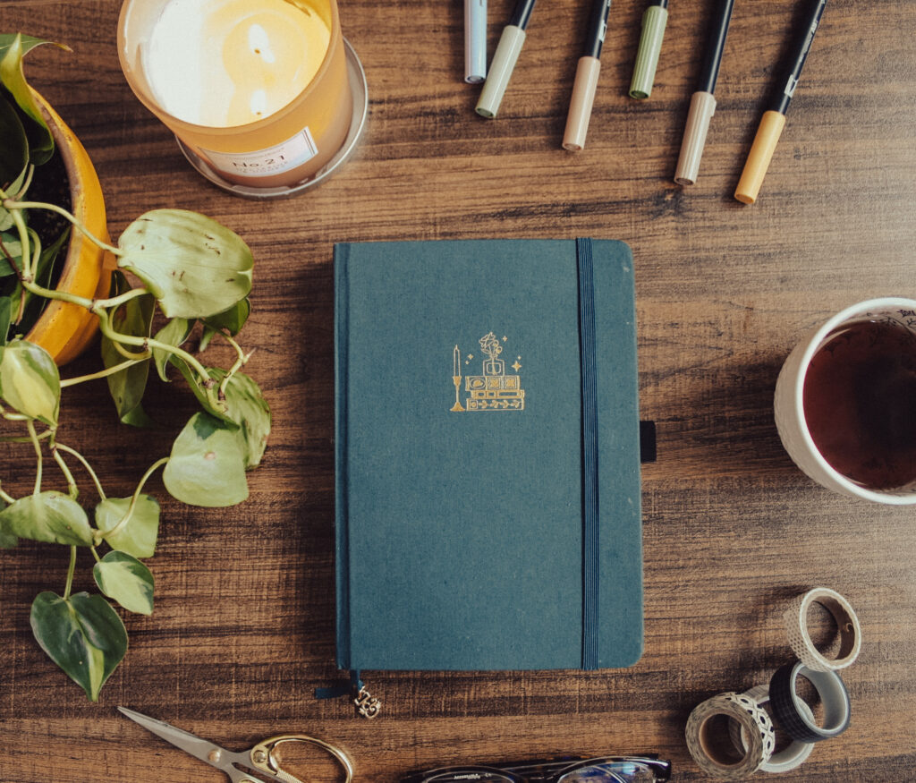 A dark blue notebook is lying on a dark surface surrounded by journaling supplies. The cover has a vintage book embossed in gold.