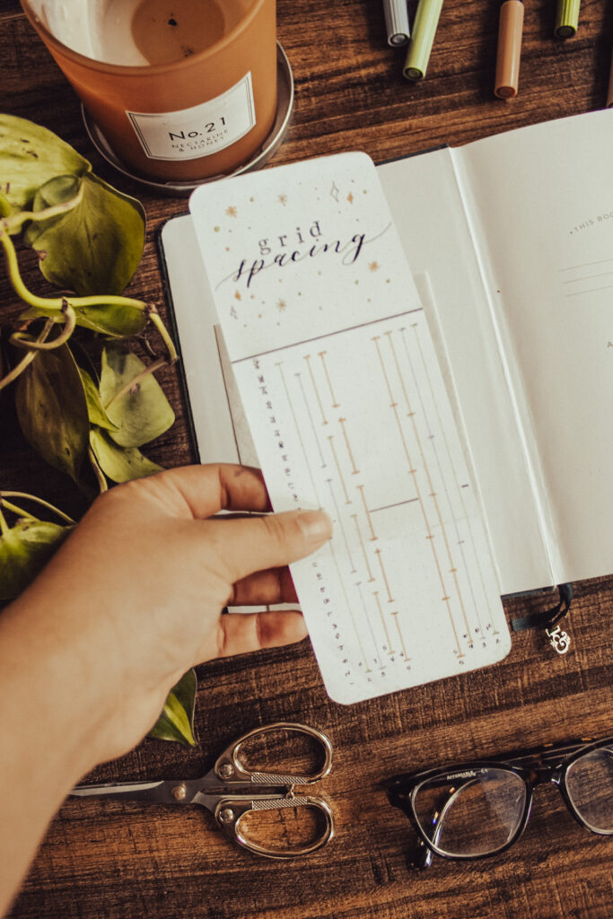 Emily is holding open a dot grid spacing guide bookmark above an open notebook. Notebook is lying on a dark table surrounded by stationery supplies.