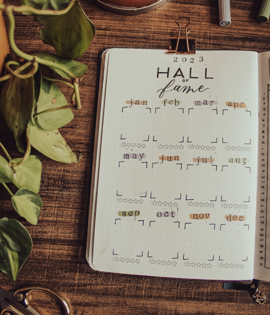 Dot grid notebook is lying open to a 2023 Books Hall of Fame spread. Square for each month where favorite book from each month will go. Notebook is lying on a dark surface surrounded by decorative elements.