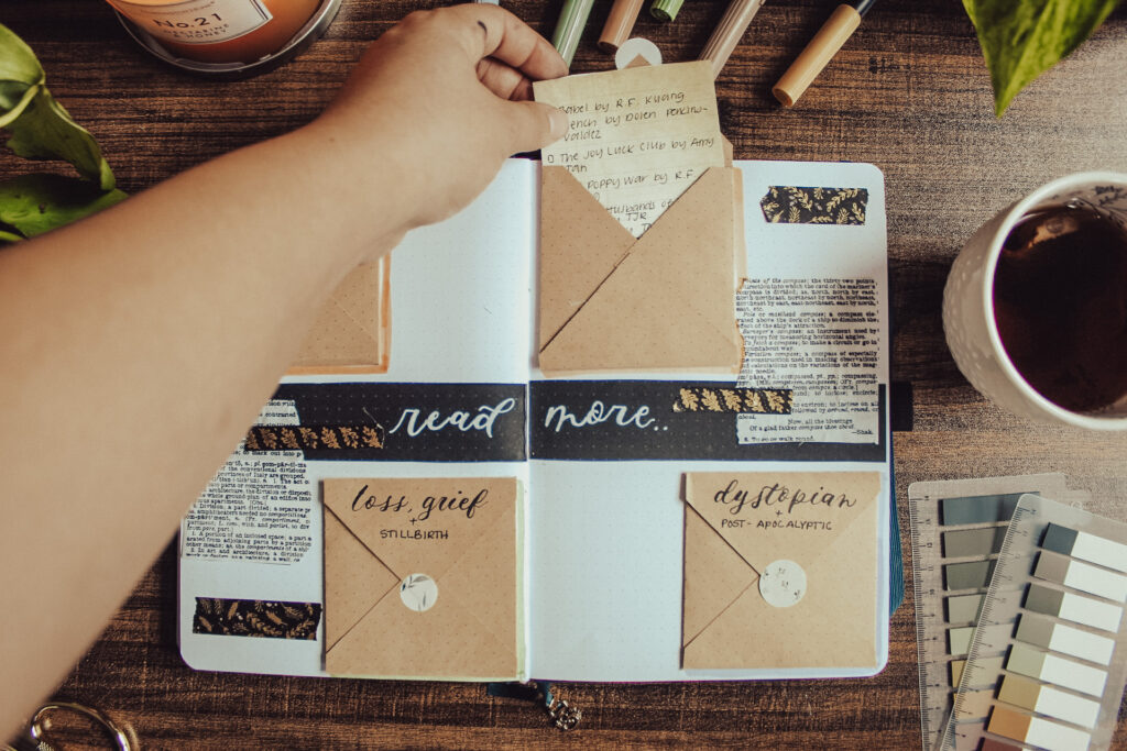 Emily is opening one of the small kraft envelopes in her genre-specific TBR spread and pulling out a list of books. Notebook is open to a book journal spread for tracking monthly stats. Notebook is lying on a dark surface surrounded by decorative elements.