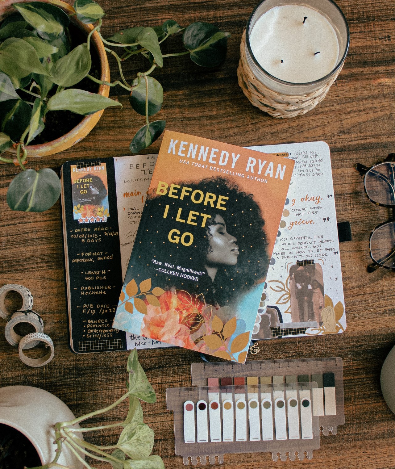 A paperback copy of the book Before I Let Go by Kennedy Ryan is on top of a reading journal spread about the book. The journal and book are on a wood desk surrounded by plants and stationery items.
