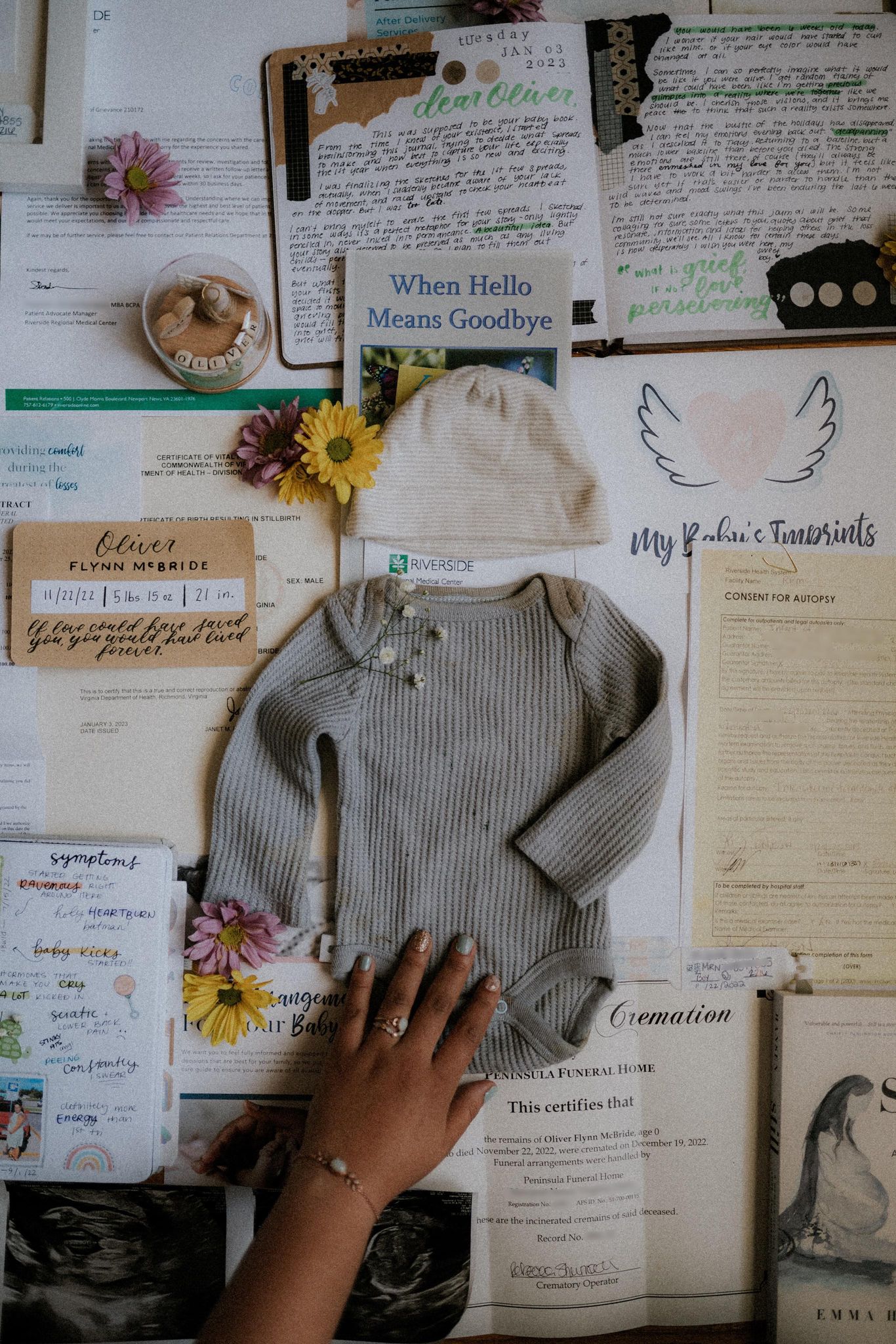 A photo highlighting grief and life after pregnancy loss, miscarriage, or stillbirth. A mother's hand with memorial jewelry has her hands on a bloodied onesies. The onesies is surrounded by various grief entries, hospital forms, books, and flowers.