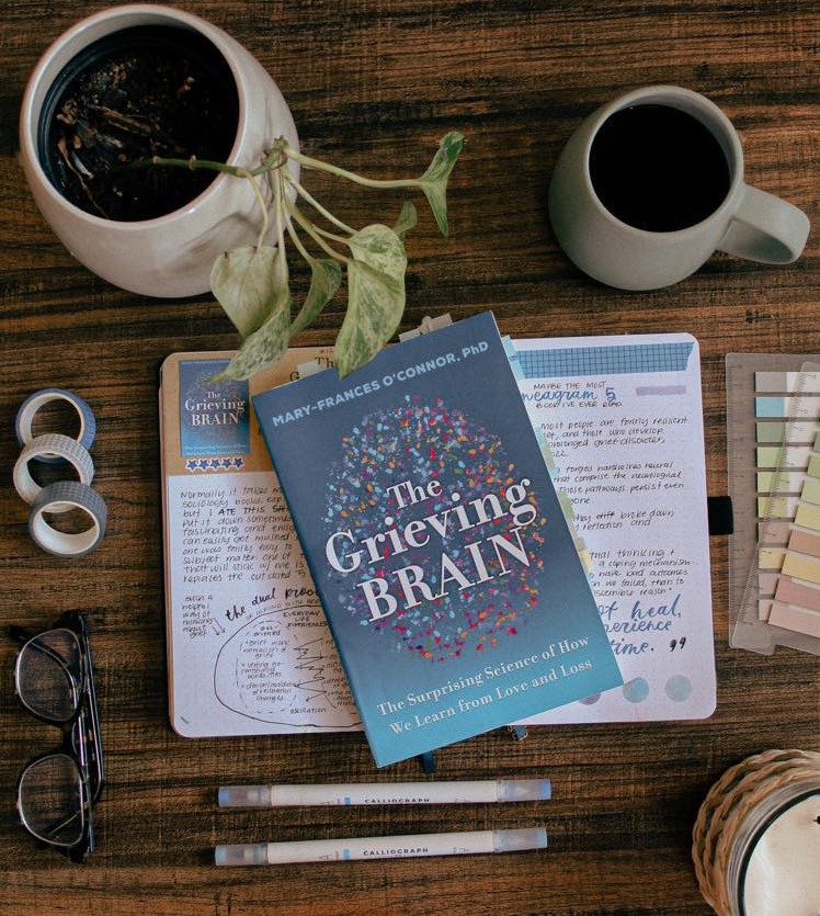 A copy of the book The Grieving Brain by Mary Frances O'Connor is laying on top of a book journal spready for the book. On the table is a plant, coffee cup, and other stationery items.