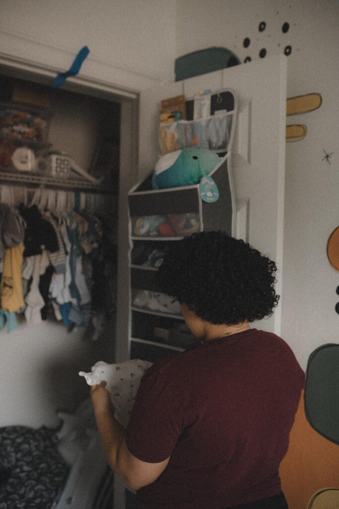 A woman is standing in front of an organized baby's closet holding a onesie in her hand. This photo is meant to highlight grief and life after miscarriage, pregnancy loss, stillbirth, or baby loss.