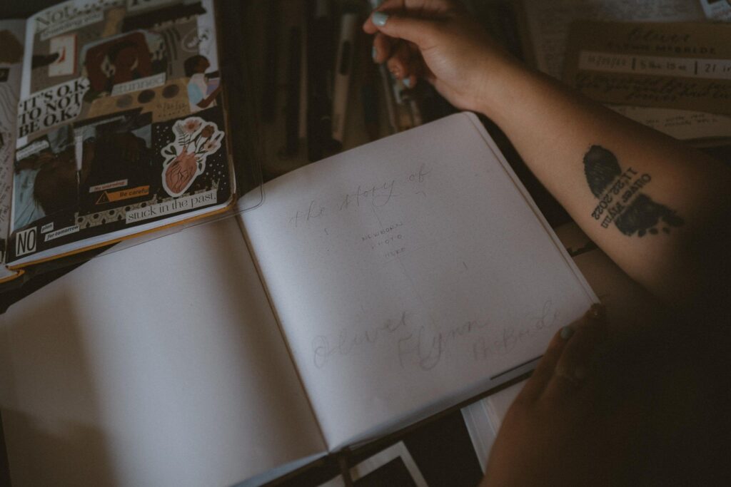 A journal is lying open with the words "The Story of Oliver Flynn McBride" sketched onto it. Emily's arm, with a memorial tattoo for her stillborn son, is lying next to the journal. Other stationery elements are on the desk.
