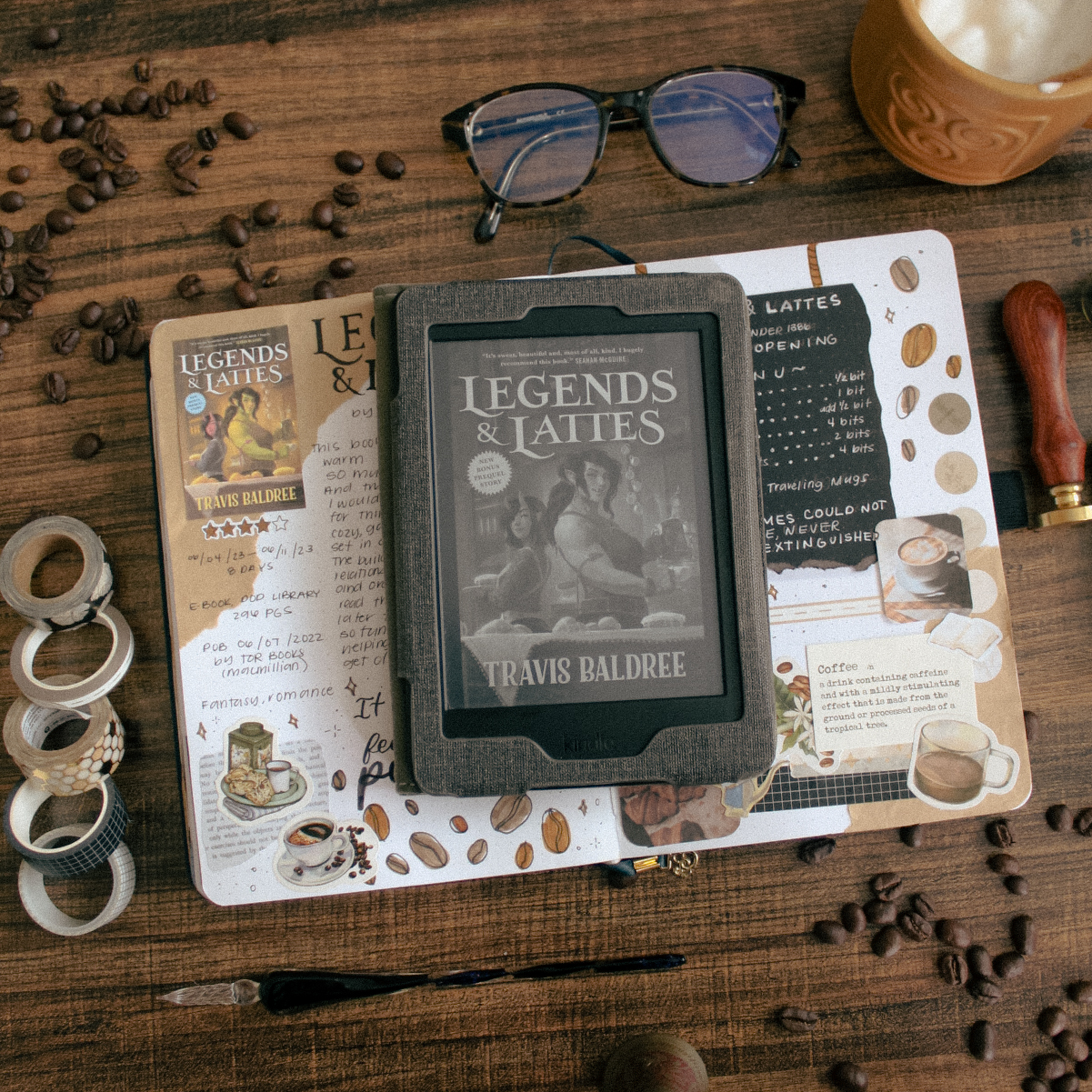 A flat-lay photo of the book Legends & Lattes by Travis Baldree. The book is lying on a reading journal book review spread for the book. On the table around it are plants and stationery elements.