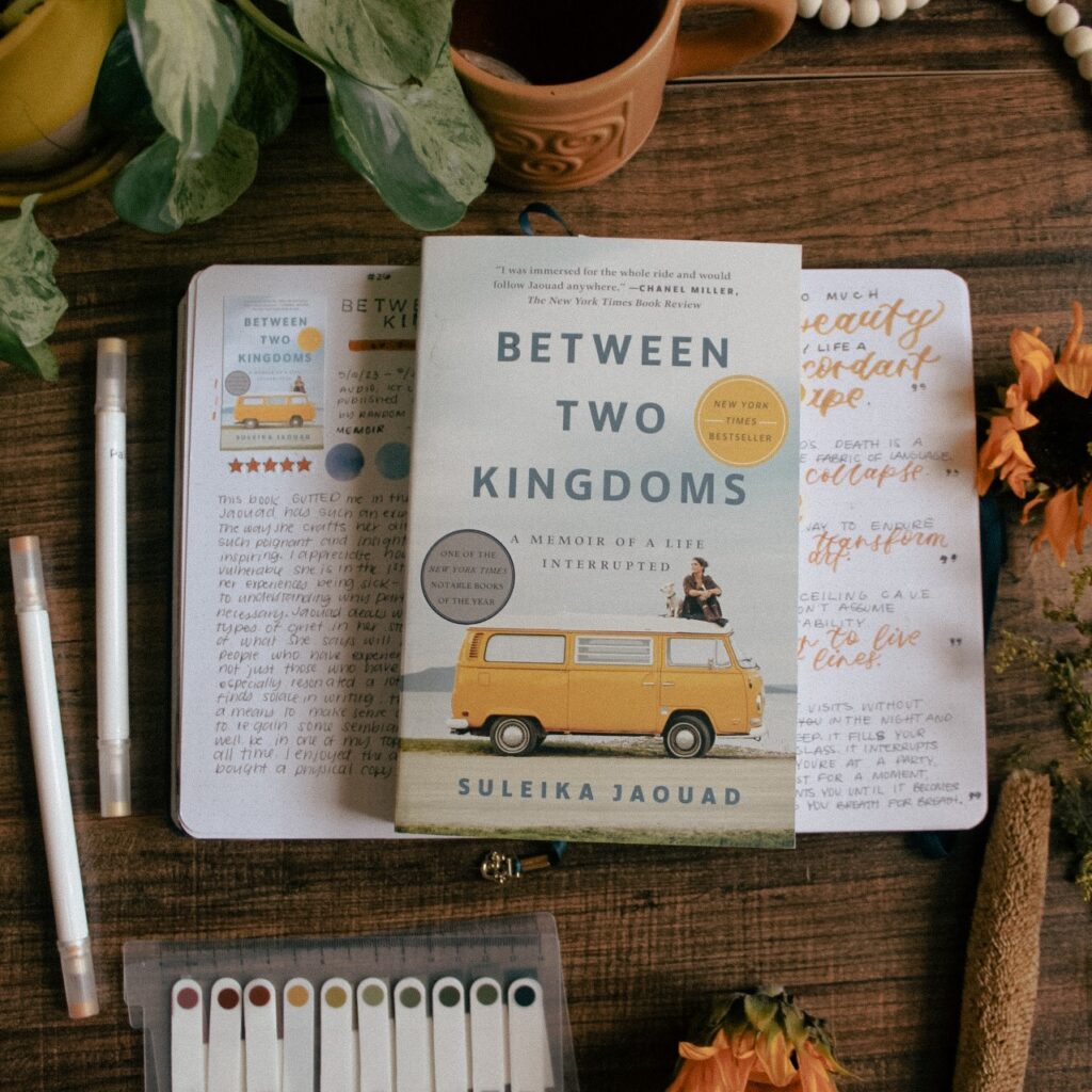 A flat-lay photo of the book Between Two Kingdoms by Suleika Jaouad. The book is lying on a reading journal book review spread for the book. On the table around it are plants and stationery elements.
