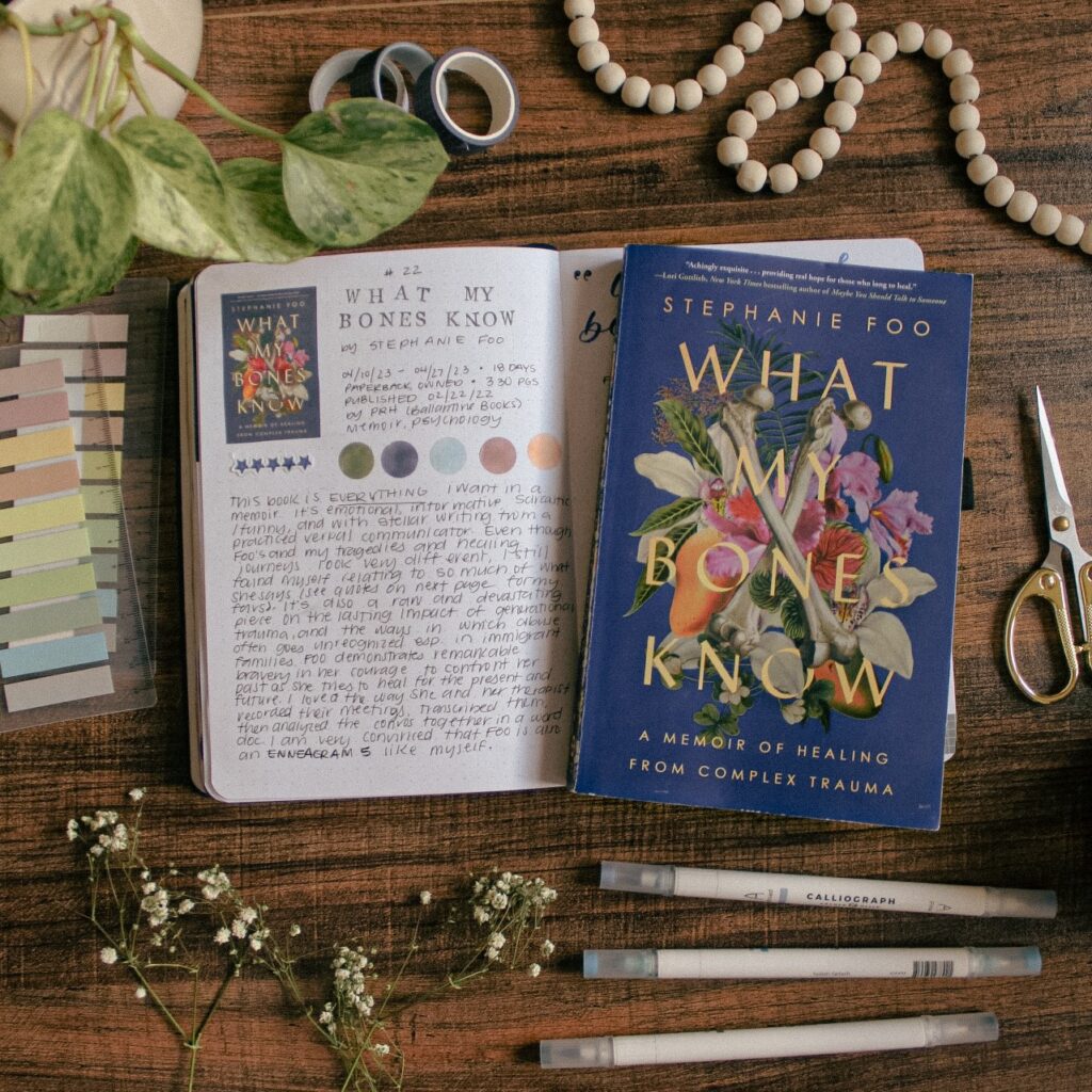A flat-lay photo of the book What My Bones Know by Stephanie Foo. The book is lying on a reading journal book review spread for the book. On the table around it are plants and stationery elements.