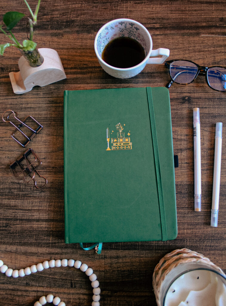 A dark green B5 sized notebook with a gold bookstack icon is sitting on a desk. On the desk around the notebooks are various decorative and stationery elements.