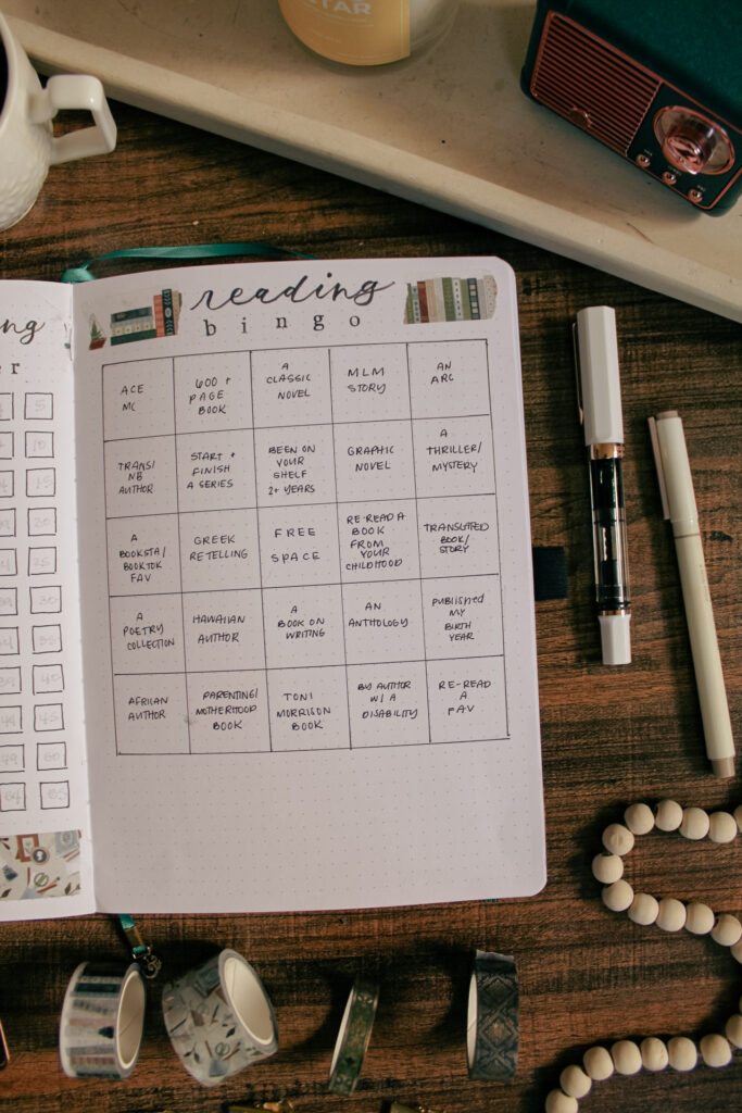 Emily's reading journal is open to a page containing her reading bingo page. On the desk around the notebook are various decorative and stationery elements.